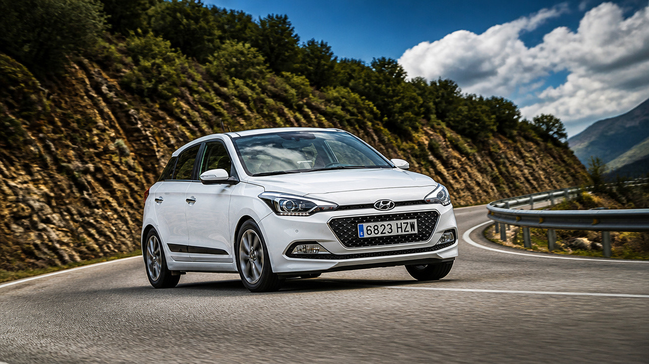 Carro branco Hyundai em curva de estrada montanhosa com guardrail e vegetação ao fundo.