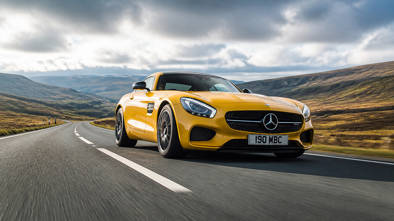 Carro esportivo amarelo Mercedes-Benz trafegando em estrada deserta entre montanhas sob céu nublado.