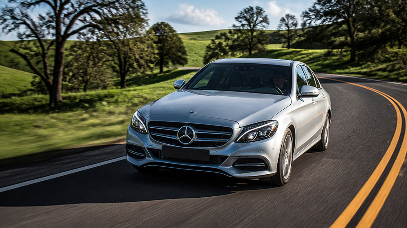 Carro prata Mercedes-Benz dirigindo em estrada sinuosa com campos verdes e árvores ao redor.
