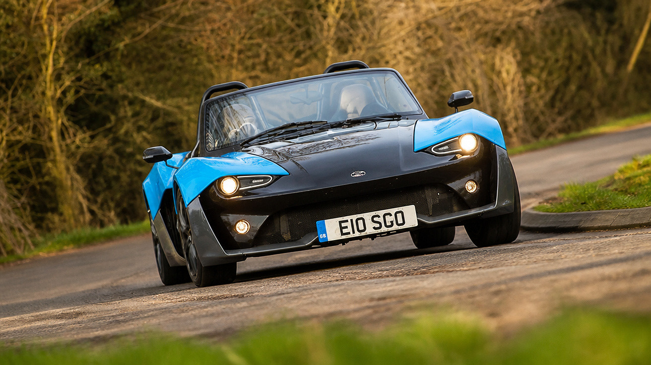 Carro esportivo conversível azul e preto dirigindo em estrada curva com vegetação ao fundo.
