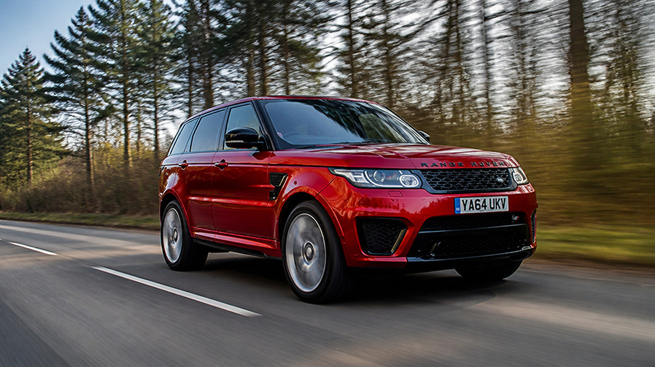 Carro SUV vermelho Range Rover trafegando em estrada cercada por árvores altas em dia claro.