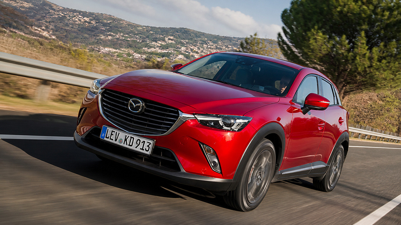 Carro vermelho Mazda CX-3 em movimento na estrada com paisagem montanhosa ao fundo.